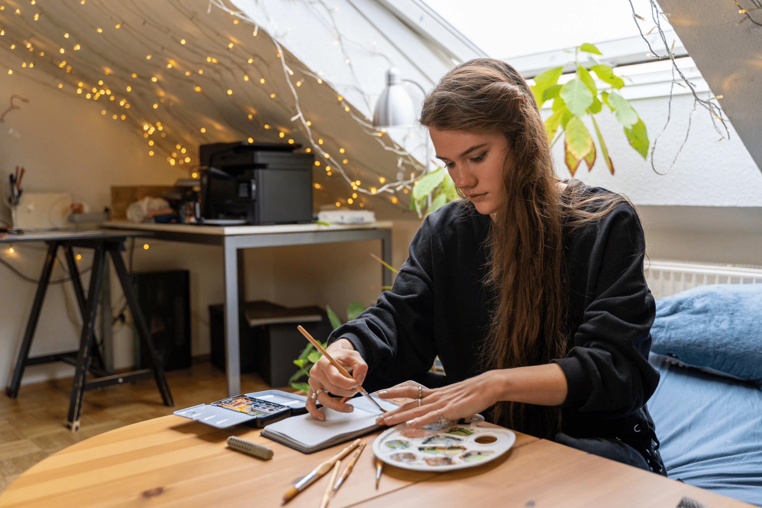 A college girl creating dorm room decorations with paints and a paintbrush, surrounding by twinkling lights