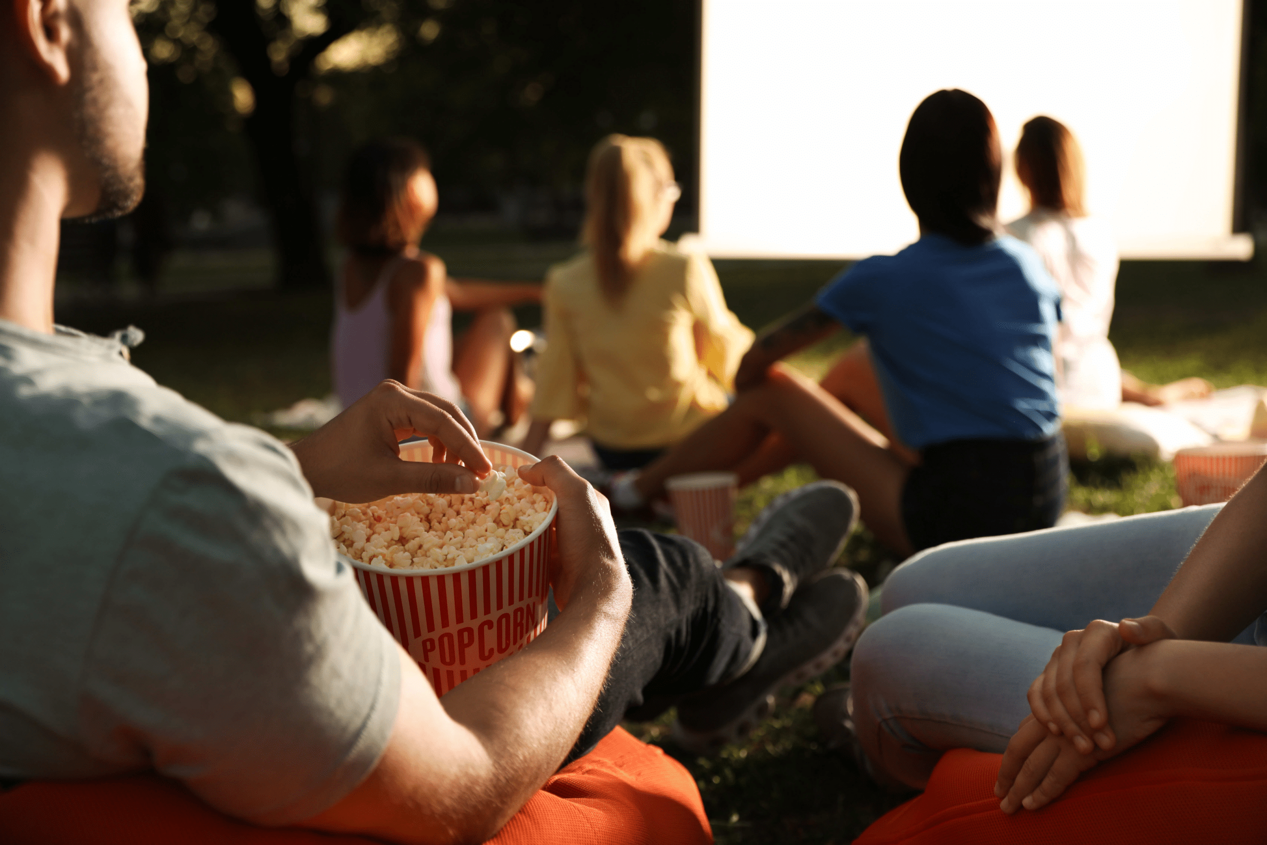 A group of college students outside sitting on the ground watching a movie on a big screen, with one guy eating popcorn