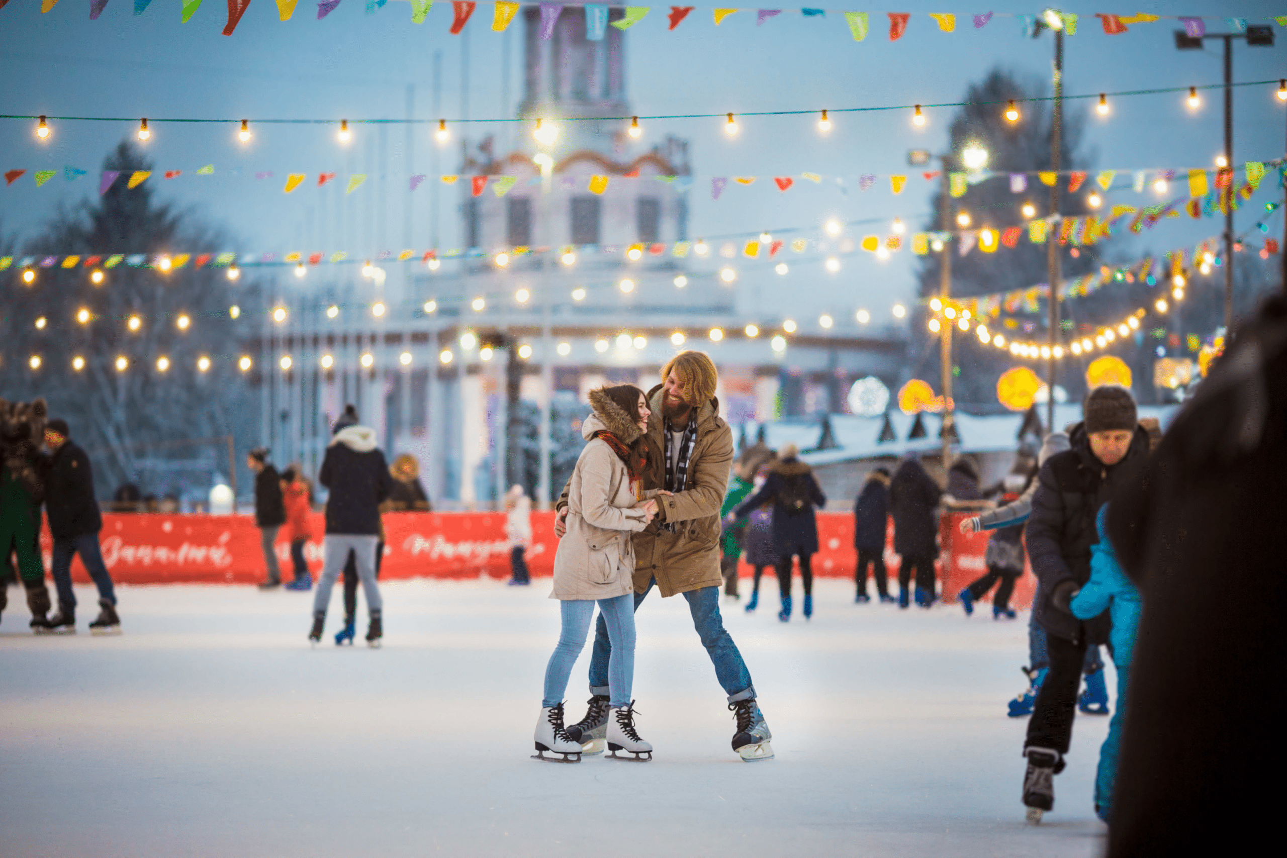 A couple skating on an ice skating rink downtown with lots of string lights and small flags above them