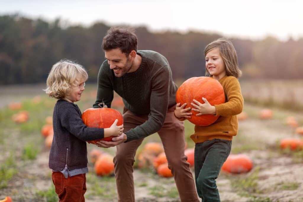Father and two kids picking pumpkins at a pumpkin patch during the fall season.