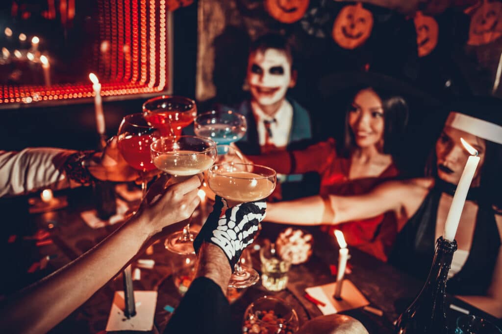 Group of Young Happy People Wearing Costumes at Halloween Gala Sitting at Table and Drinking Cocktails.