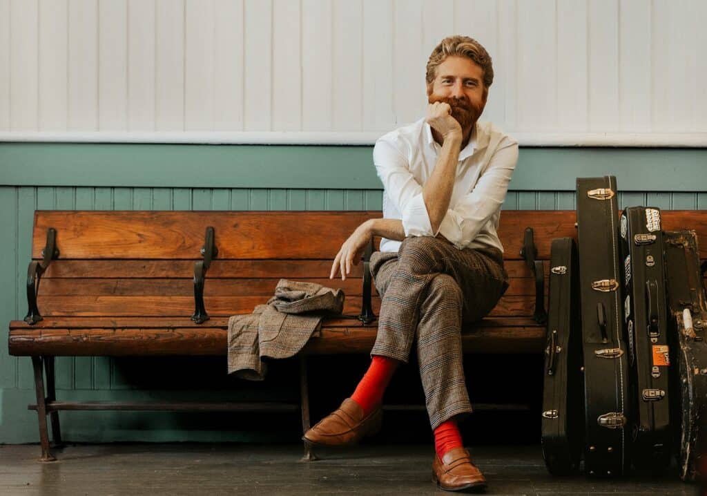 sean dietrich sitting at a train station bench with his guitar cases