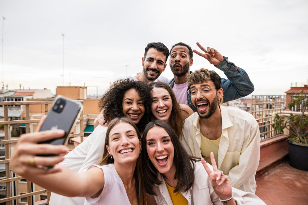 Diverse joyful group of carefree friends having fun and taking a selfie at a rooftop party. Multiracial beautiful young happy people laughing while taking a picture at a terrace.