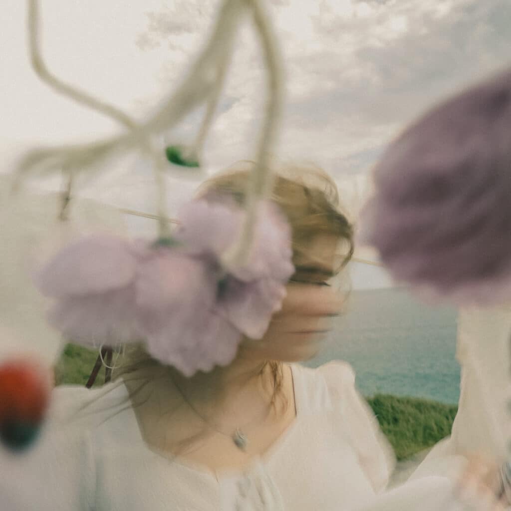 image of Marina Anthea in a white dress surrounded by flowers with her face artistically blurred as she moves it